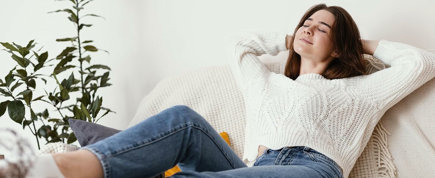 Mujer relajándose en el sofá vistiendo un suéter de punto blanco y jeans azules. Taza de café y planta visibles en primer plano. Acogedor entorno hogareño con iluminación tenue.