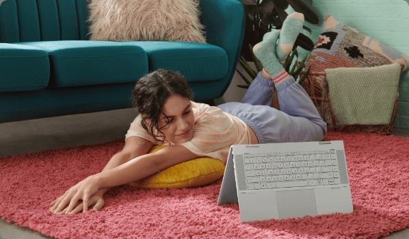 امرأة شابة مستلقية على الأرض تنظر إلى كمبيوتر محمول HP ENVY x360 في وضع الخيمة
